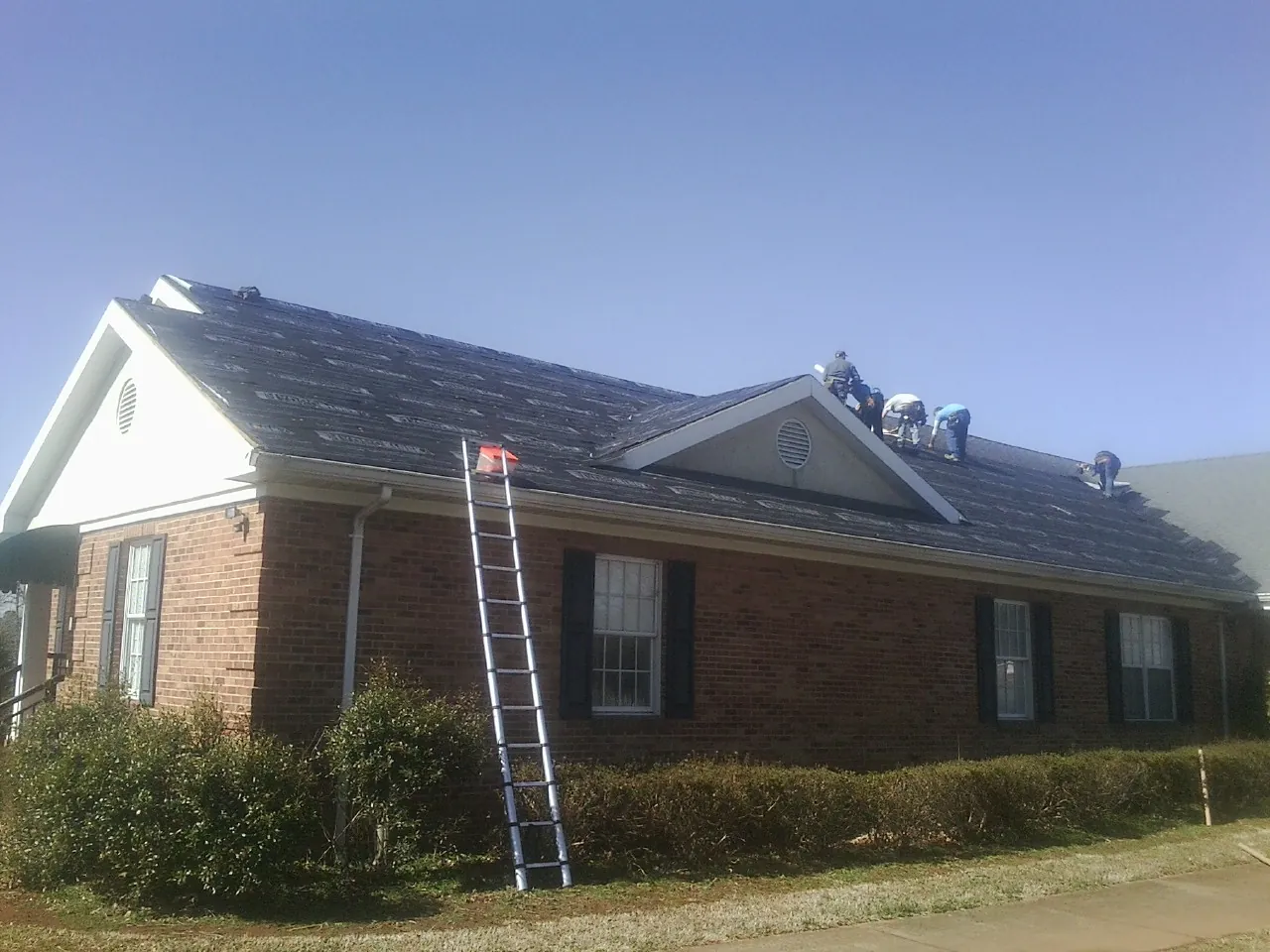 Crew tearing shingles off a brick ranch roof with a ladder set — Emergency Roof Repair work by our crew in Belews Creek