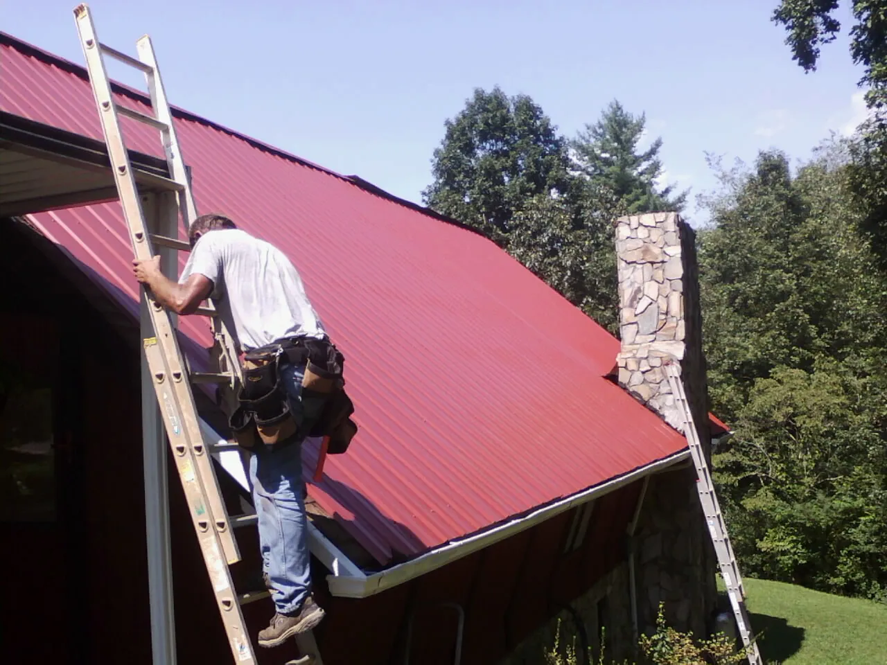 Roofer on a ladder beside a red metal roof and stone chimney — Soffit & Fascia Repair by Roofing Brothers in Belews Creek